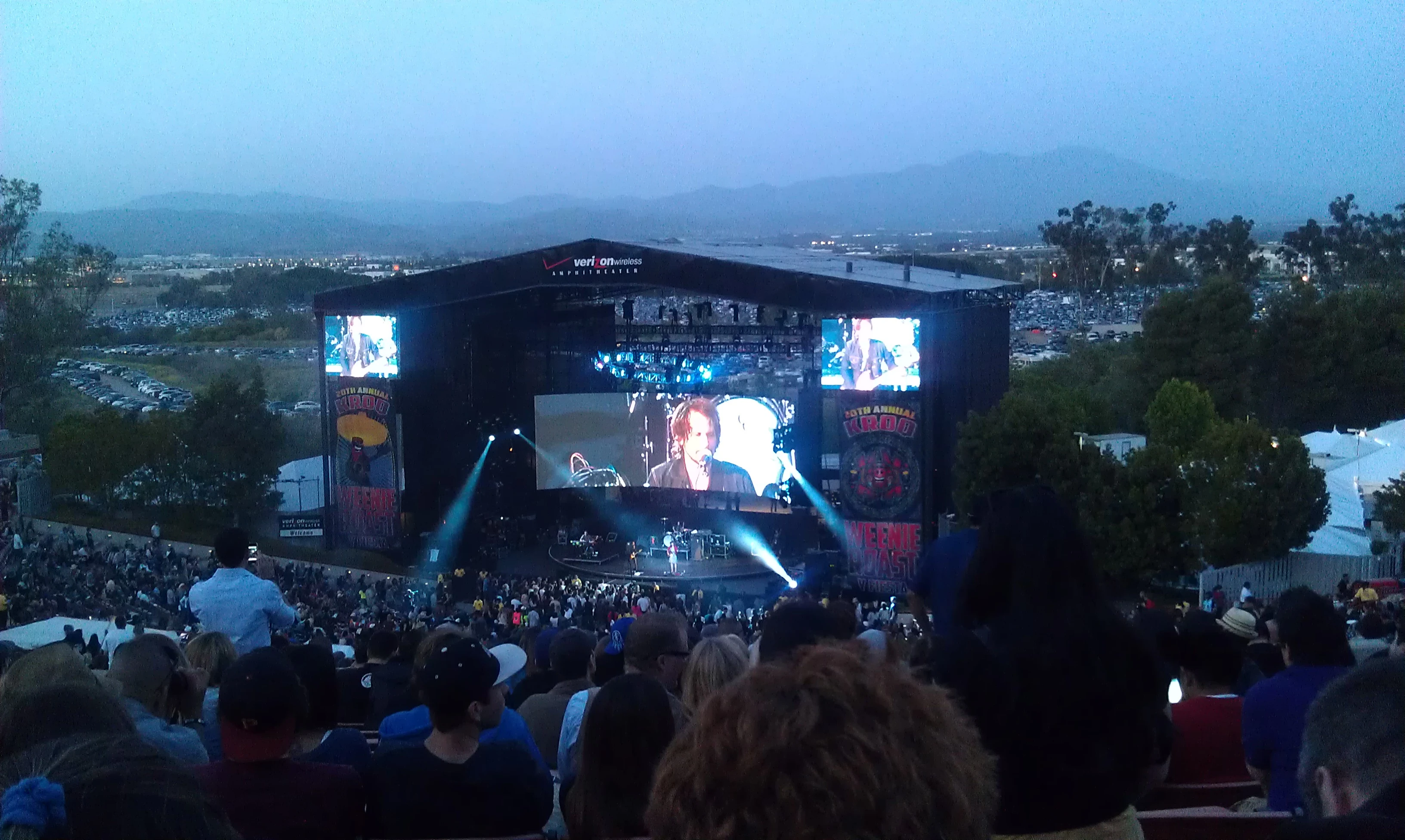 Outdoor amphitheater concert at dusk, a large stage with blue spotlights and a giant LED screen showing a close-up of a performer's face, mountains visible on the horizon, a packed crowd filling the venue