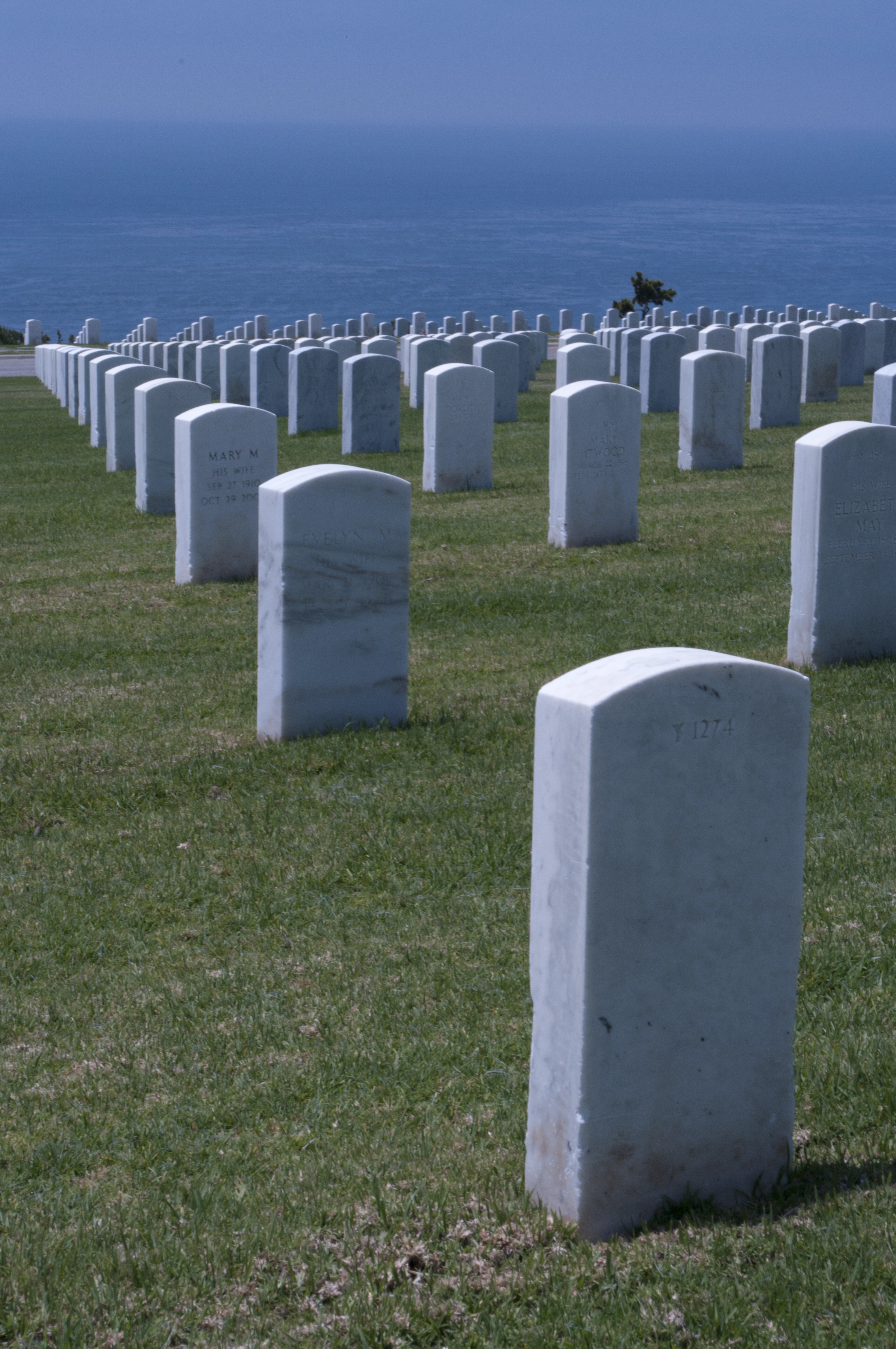 White military headstones spread across a gently sloping green lawn at Fort Rosecrans National Cemetery, with the deep blue Pacific Ocean visible on the horizon