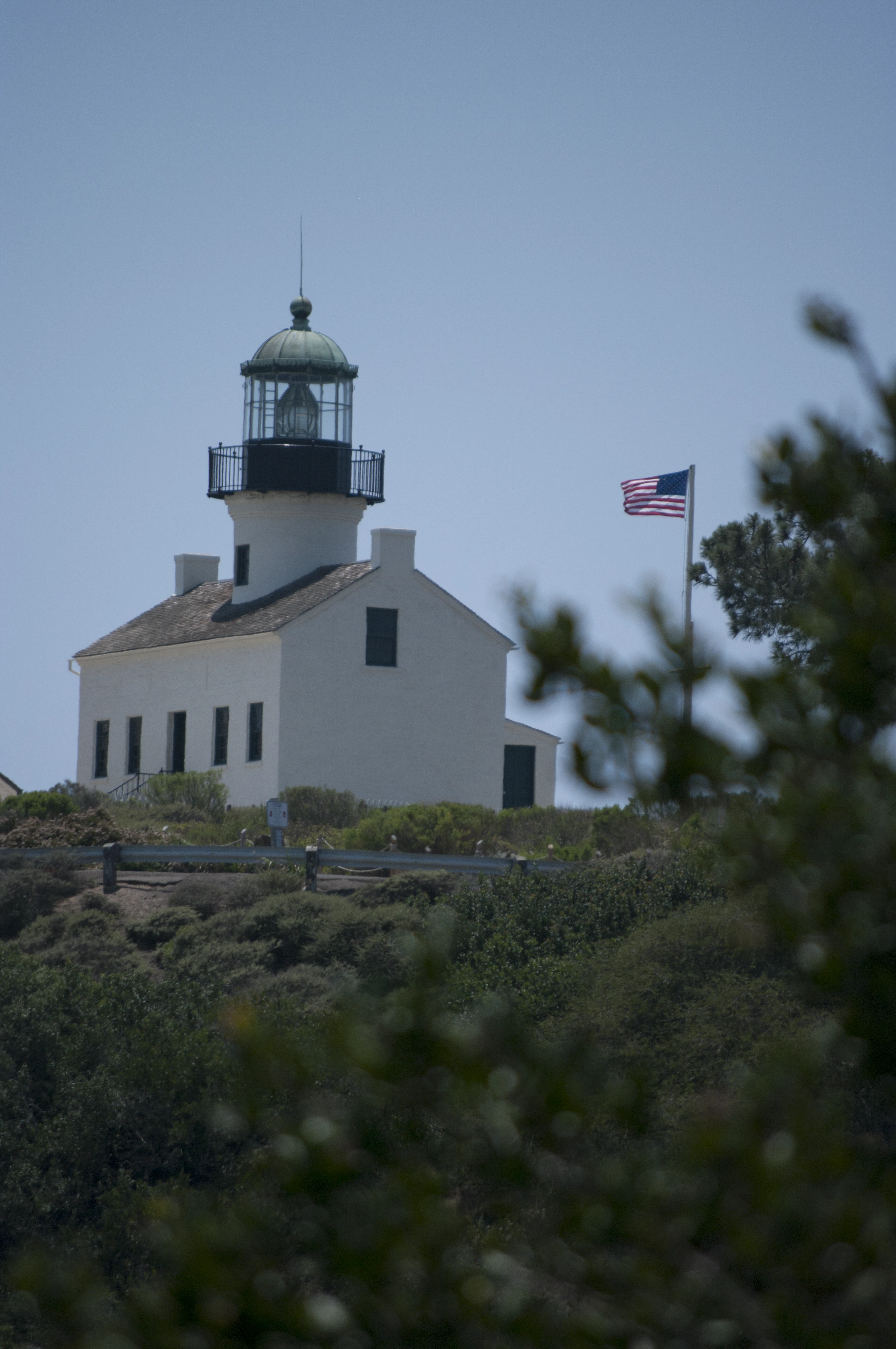 The Old Point Loma Lighthouse seen from a distance through tree branches, its white walls and green-domed tower rising above a hilltop, an American flag flying to its right against a grey-blue sky
