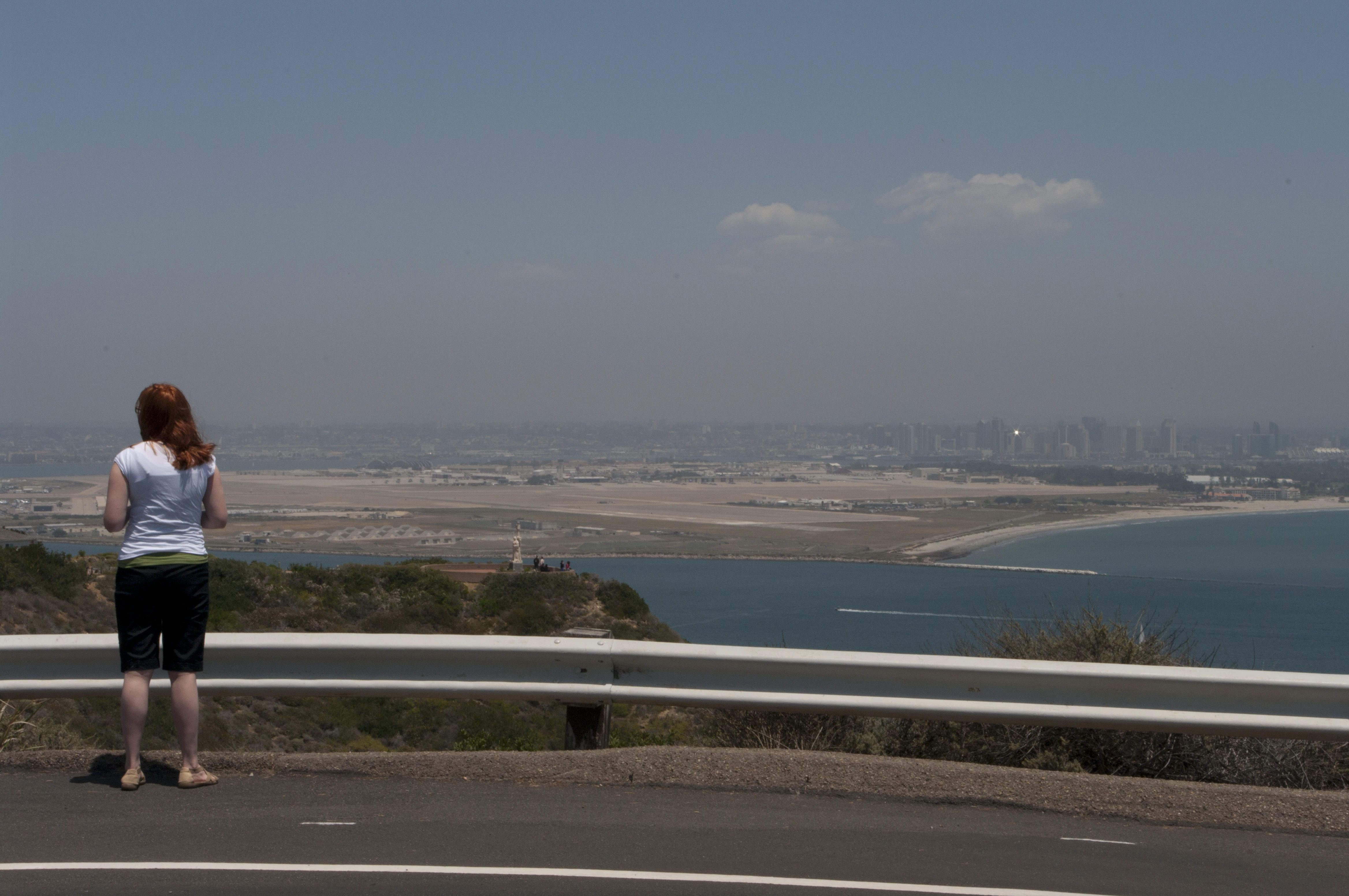 Danielle standing at a guardrail on a hillside road, looking out over a sweeping view of San Diego Bay, the airport, and the hazy downtown skyline in the distance